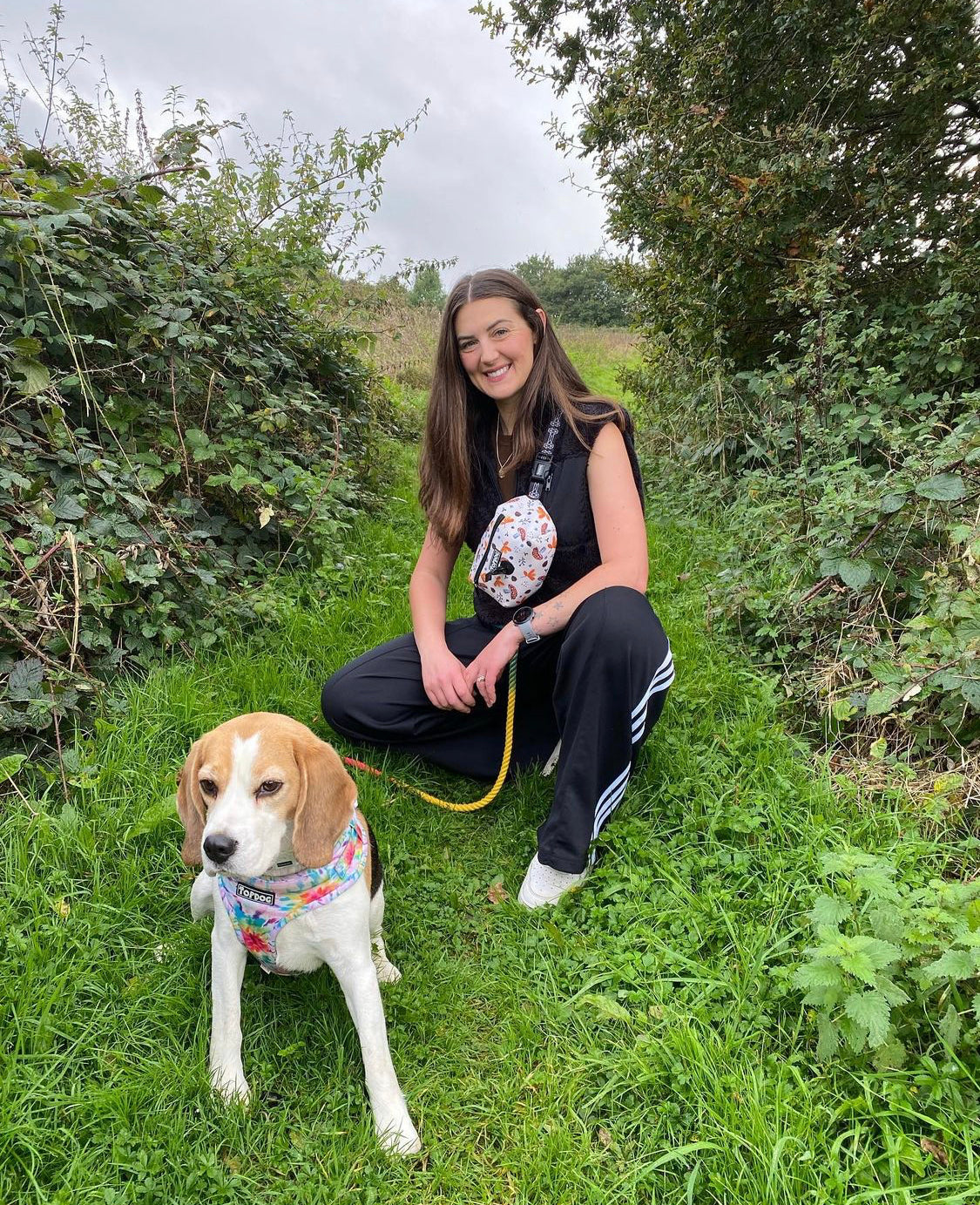 Beagle with young woman owner wearing topDog Harnesses Matching Essential Dog walking bag, sitting on grass on a country walk