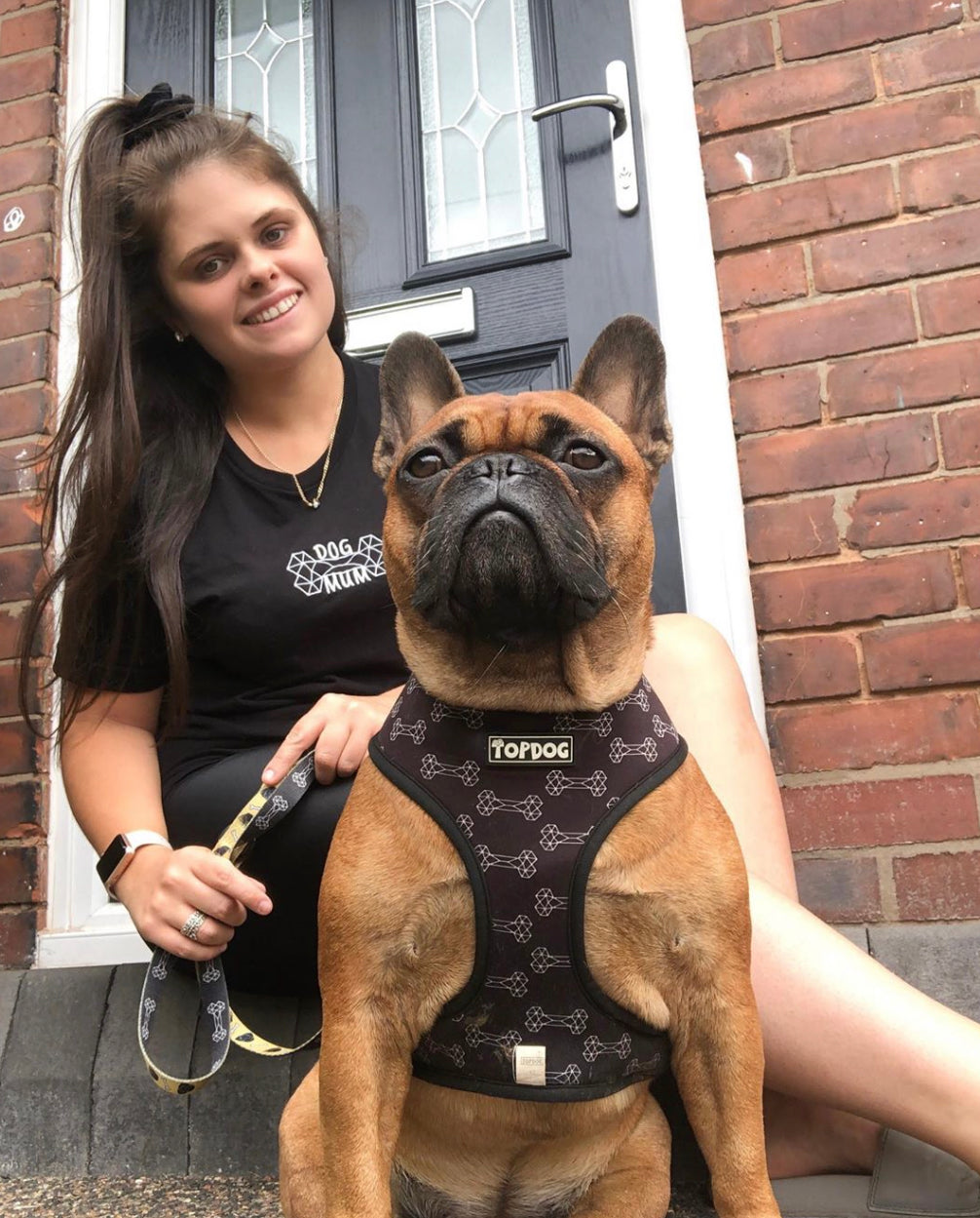 French Bulldog wearing a TopDog Harnesses Give a Dog a Bone Reversible dog harness with young woman owner in front of their door 